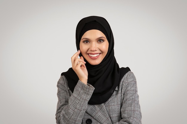 Portret muzułmańskiej bizneswoman w hidżabie uśmiechającej się do kamery elegancko pozującej na tle szarego studia