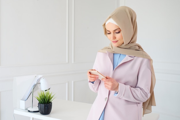 Portret muzułmańskiej kobiety w beżowym hidżabie czyta coś na telefonie komórkowym