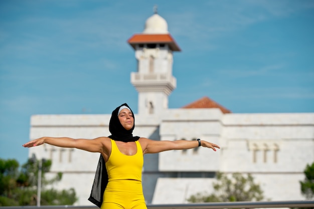 Portret Muzułmańskiej Kobiety W Sportowej Z Hidżabu Uczucie Wolności. Poziomy Widok Z Boku Fitness Arabska Kobieta Z Szeroko Otwartymi Ramionami Na Białym Tle W Tle Meczetu.