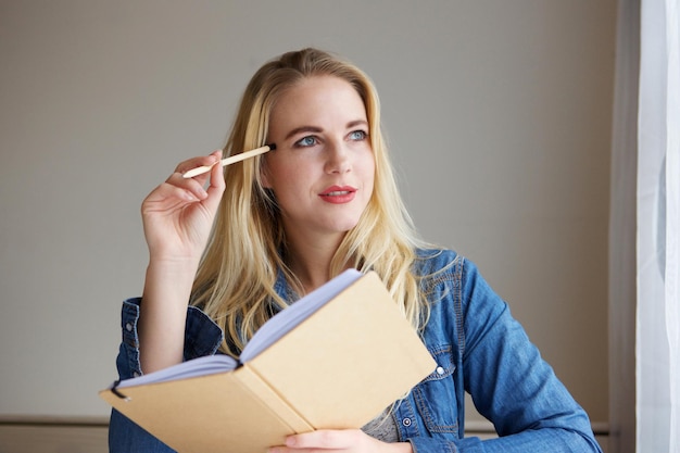 Portret myślącej blondynki trzymającej książkę i ołówek