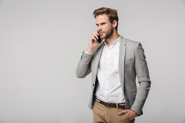 Portret myślący przystojny mężczyzna w kurtce opowiada na telefonie komórkowym odizolowywającym nad białą ścianą