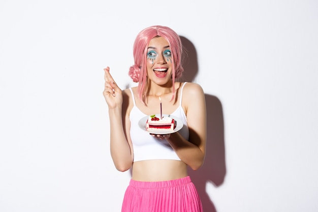 Portret nadziei urodzinowej dziewczyny w różowej peruce, życzącej ze skrzyżowanymi palcami, trzymającej tort urodzinowy, stojącej na białym tle