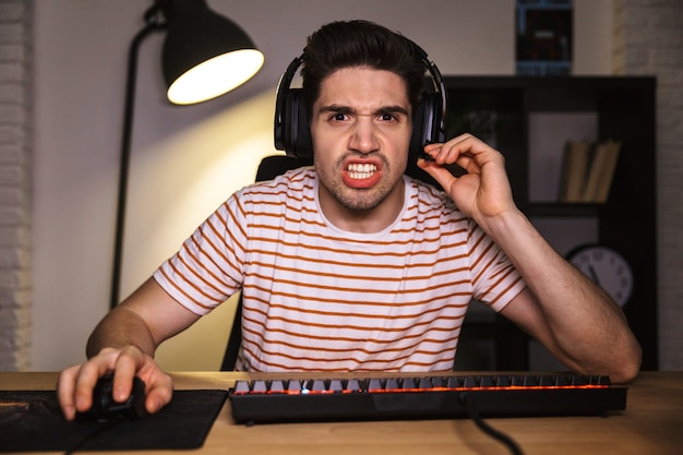 Portret napiętego młodzieńca 20s noszenie zestawu słuchawkowego krzycząc, siedząc przy biurku z komputerem w pokoju i patrząc na monitor