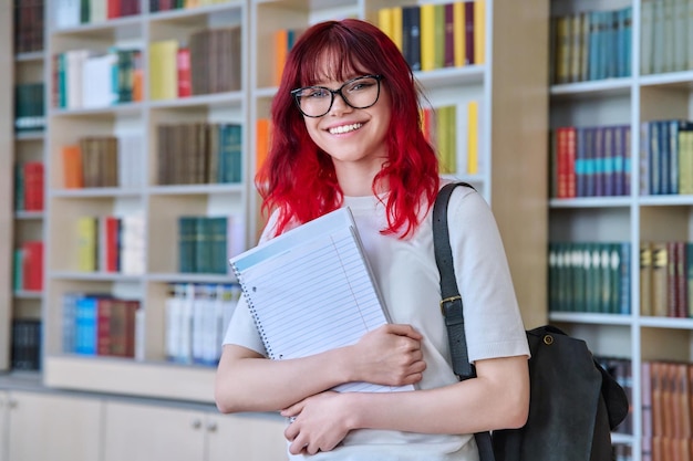 Portret nastoletniej studentki patrzącej na kamerę w bibliotece Uśmiechnięta piękna nastolatka w okularach trzymająca plecak na notebooka Koncepcja młodości wiedzy o edukacji uniwersyteckiej
