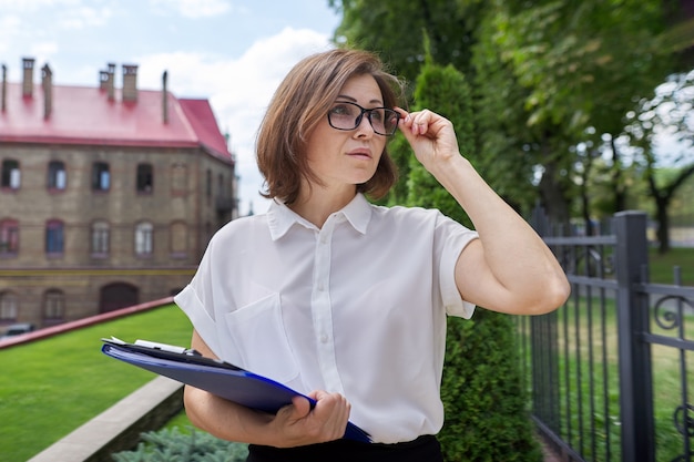 Portret nauczyciela dojrzałych businesswoman, menedżera z dokumentów biznesowych dokumentów w ręce. Budynek szkoły, tło budynku biurowego, profesjonalna pewna siebie kobieta w okularach biała koszula