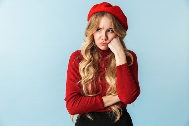 Portret niezadowolona blond kobieta 20s ubrana w czerwony beret, patrząc na bok stojąc, na białym tle