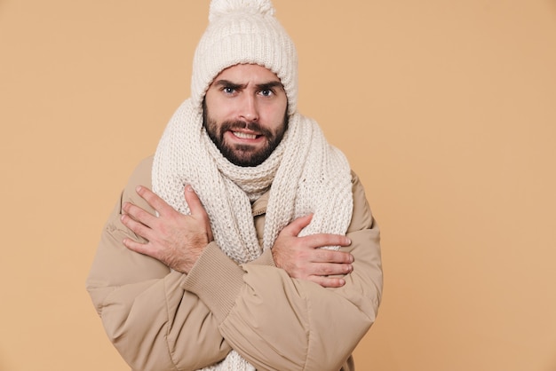 Portret niezadowolonego młodego mężczyzny w zimowych ubraniach drży i drży z zimna na beżowym tle