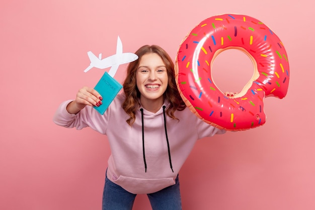 Portret niezwykle szczęśliwej kręconej nastoletniej dziewczyny w bluzie z kapturem z zębatym uśmiechem, trzymającej gumowy pierścień pączka, paszport i papierowy samolot. Kryty strzał studio na białym tle na różowym tle