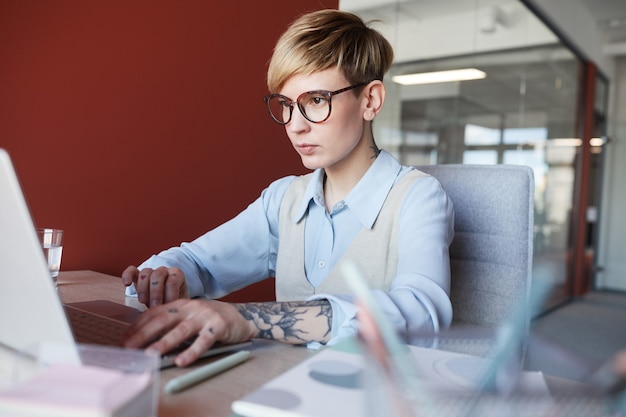 Zdjęcie portret nowoczesnego wytatuowanym businesswoman za pomocą laptopa przy biurku podczas pracy w biurze przed czerwoną ścianą, kopia przestrzeń