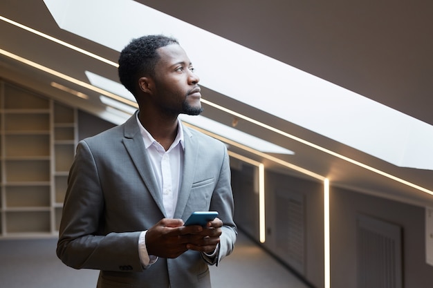 Portret Odnoszącego Sukcesy Afrykańsko-amerykańskiego Biznesmena Trzymającego Smartfona I Odwracającego Wzrok W Zadumie, Stojąc W Minimalistycznym Szarym Wnętrzu Nowoczesnego Biura Oświetlonego Przez świetliki, Kopiuj Przestrzeń