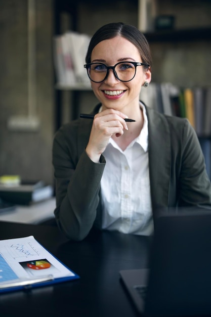 Portret odnoszącej sukcesy młodej bizneswoman siedzącej przy biurku pracującej na komputerze przenośnym uśmiechającej się do kamery