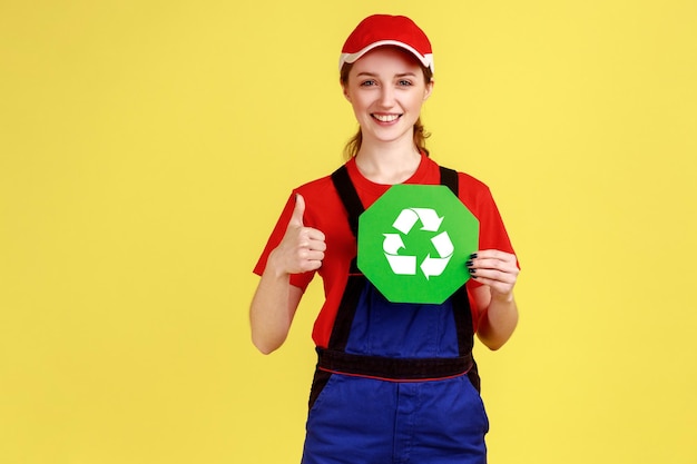 Portret optymistycznego pracownika kobiety stojącej i trzymającej znak recyklingu myślący zielony pokazując kciuk do góry ubrany w kombinezon i czerwoną czapkę Kryty studio strzał na białym tle na żółtym tle