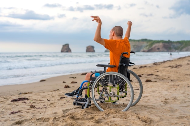 Portret osoby niepełnosprawnej na plecach w wózku inwalidzkim na plaży z otwartymi ramionami cieszący się wolnością morza i przyrody