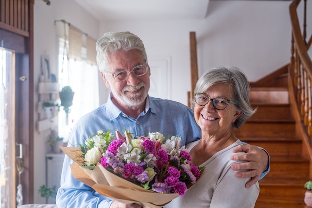 Portret para dwóch szczęśliwych i zakochanych seniorów lub dojrzałych i starych ludzi posiadających kwiaty w domu patrząc w kamerę. Emeryci i renciści cieszą się i wspólnie świętują wakacje.