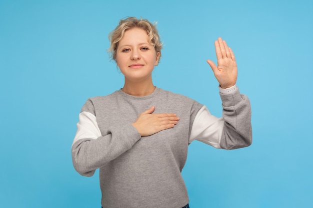 Portret patriotycznej obywatelki, uczciwej dorosłej kobiety z kręconymi włosami w bluzie trzymającej rękę na klatce piersiowej i unoszącej dłoń, składającej przyrzeczenie lojalności, przysięgę. strzał w studio na białym tle na niebieskim tle