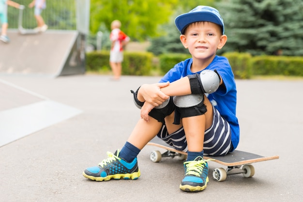 Portret pełnej długości uśmiechający się młody chłopak noszący ochronne ochraniacze na łokcie i kolana, siedząc na deskorolce w odkrytym skateparku