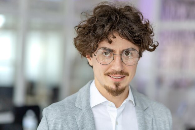 Portret pewnego siebie młodego biznesmena z kręconymi włosami noszącego okulary i uśmiechającego się w jasny