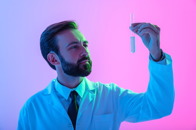 Portret pewnego siebie naukowca młodego człowieka w płaszczu, stojącego na białym tle nad różowo-niebieską mgiełką, patrzącego na rurkę medyczną z płynem