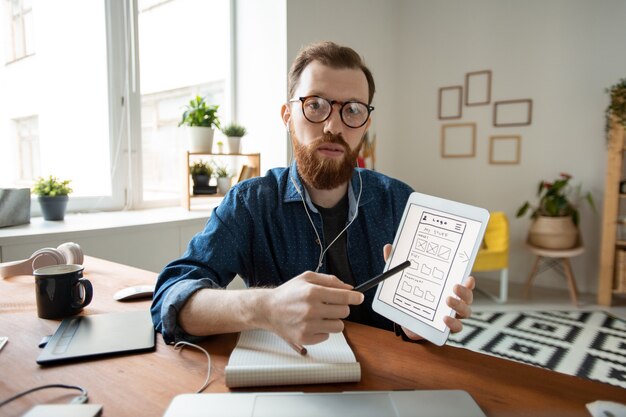 Zdjęcie portret pewnego siebie projektanta interfejsu użytkownika w okularach, wskazującego na ekran tabletu i prezentującego projekt interfejsu podczas rozmowy przez aplikację do wideokonferencji