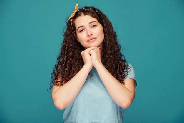 Portret pewnej siebie dziewczyny z kręconymi włosami w crop top, trzymając się za ręce na biodrach na tle niebieskiej ściany