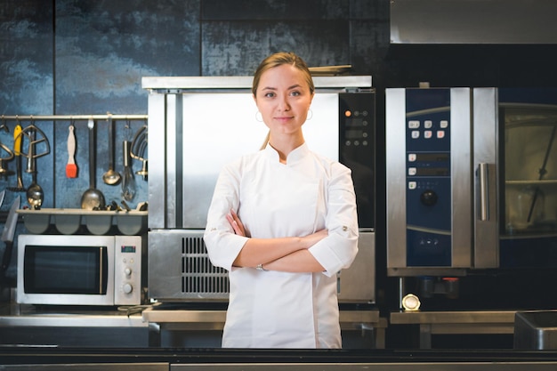 Portret pewnej siebie i uśmiechniętej młodej kobiety szefa kuchni, ubranej w białą, jednolitą profesjonalną kuchnię, jest na tle