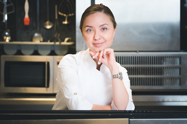 Portret pewnej siebie i uśmiechniętej młodej kobiety, szefa kuchni, ubranej w białą, jednolitą profesjonalną kuchnię, jest na tle