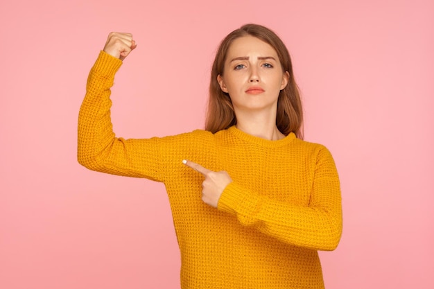 Portret pewnej siebie rudej dziewczyny w swetrze, wskazując na jej biceps pokazujący moc w dłoni i patrzący dumnie czując silny i niezależny feminizm w pomieszczeniu studio strzał na białym tle na różowym tle