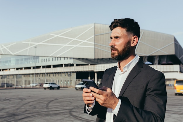 Portret pewny siebie biznesmen w klasycznym garniturze podczas rozmowy wideo z partnerem biznesowym przy użyciu telefonu komórkowego stojącego w pobliżu nowoczesnego biurowca i białego samochodu. Koncepcja technologii.