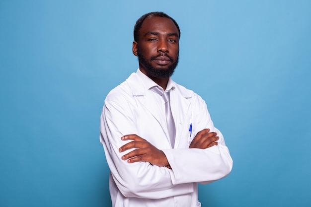 Portret pewny siebie lekarz w białym fartuchu na niebieskim tle w studio fotografii. Medyk w mundurze laboratoryjnym pozuje ze skrzyżowanymi rękami. Osoba z personelu medycznego.