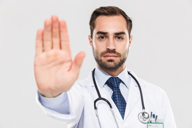 Portret pewny siebie młodego lekarza ze stetoskopem ściśle i pokazujący jego dłoń odizolowaną nad białą ścianą