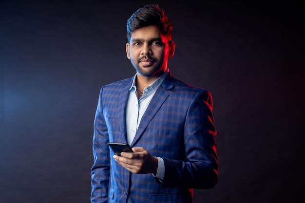 Portret pewny siebie stylowy młody indianin w formalwear, trzymając telefon komórkowy, patrząc na ekran, stojąc na czarnym tle. Sukcesy biznesmen. Biznes, technologia.