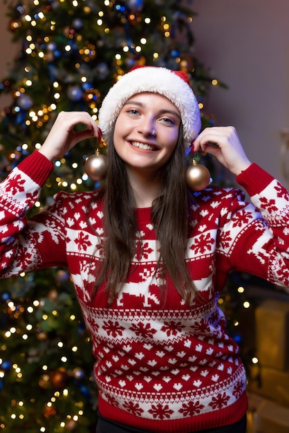 Portret piękna dziewczyna w świątecznym swetrze i czerwonym kapeluszu Świętego Mikołaja siedzi w pobliżu luksusowej choinki Uśmiechnięta trzyma ozdoby świąteczne w pobliżu głowy Obchody nowego roku bożego narodzenia