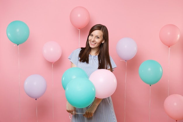 Portret piękna uśmiechnięta młoda szczęśliwa brunetka ubrana w niebieską sukienkę, trzymająca kolorowe balony na jasnym modnym różowym tle. Urodziny wakacje, koncepcja ludzie szczere emocje.