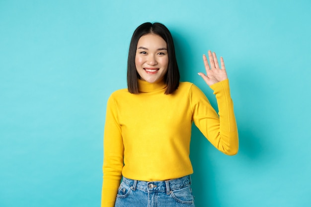Portret Pięknej Azjatyckiej Dziewczyny Ze Szczęśliwym Uśmiechem, Machając Ręką, Aby Się Przywitać, Witając Cię Przyjazną Twarzą, Stojąc Na Niebieskim Tle