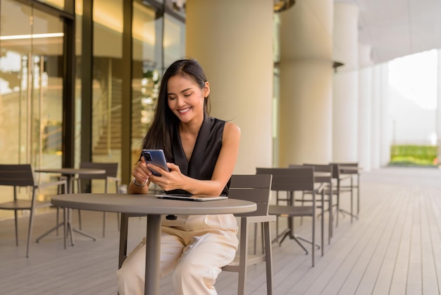 Portret pięknej azjatyckiej kobiety siedzącej na zewnątrz w kawiarni restauracji uśmiechniętej i używającej telefonu