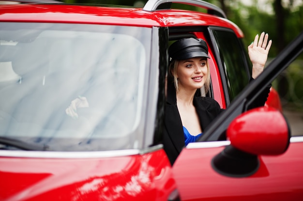 Portret pięknej blondynki seksowna kobieta modelka w czapce i we wszystkich czarnych, niebieskich gorsetach, z jasnym makijażem w pobliżu czerwonego samochodu miejskiego.