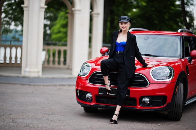 Portret pięknej blondynki seksowna kobieta modelka w czapce iw całej czerni z jasnym makijażem siedzieć na masce czerwonego samochodu miejskiego.