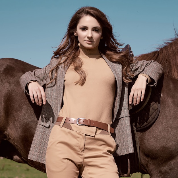 Portret pięknej brunetki kobiety w eleganckiej brązowej kurtce w kratkę z koniem na wiejskim krajobrazie