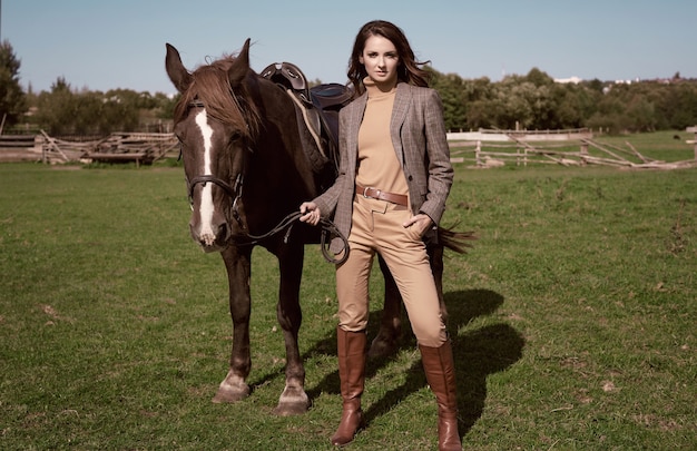 Portret pięknej brunetki kobiety w eleganckiej brązowej kurtce w kratkę z koniem na wiejskim krajobrazie