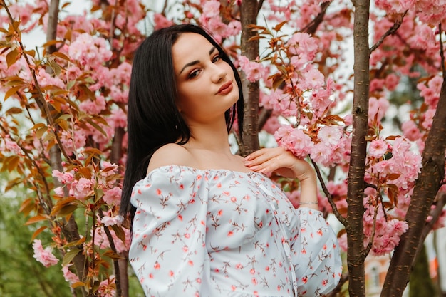 Portret pięknej delikatnej kobiety ubranej w różową kwiecistą sukienkę pozowanie w pobliżu kwiatów wiśni sakura