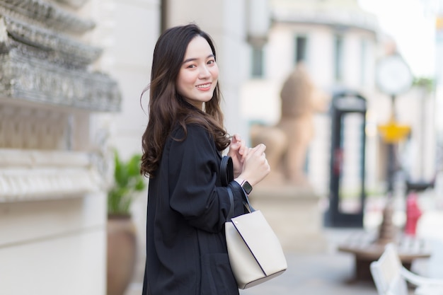 Portret pięknej, długowłosej Azjatki w czarnym płaszczu, niosącej uśmiechniętą torbę spacerową