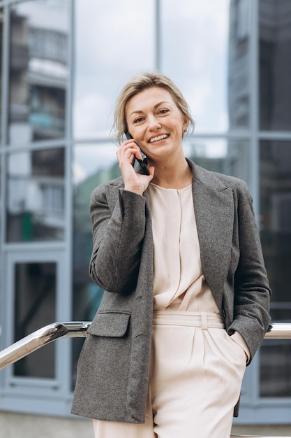 Portret pięknej dojrzałej kobiety biznesu w garniturze i szarej kurtce uśmiecha się i rozmawia przez telefon na tle nowoczesnych budynków miejskich i biurowych