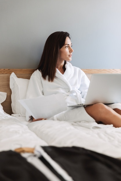 Portret pięknej dorosłej bizneswoman w białym szlafroku, pracującej z papierowymi dokumentami i laptopem, leżąc na łóżku w mieszkaniu