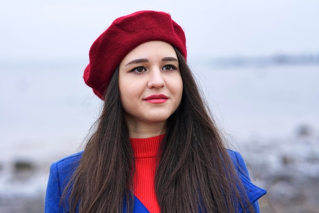 Portret pięknej dziewczyny rasy kaukaskiej, młoda atrakcyjna stylowa elegancka brunetka kobieta z długimi włosami w czerwonym berecie i rękawiczkach i niebieskim płaszczu, patrząc na kamery na zewnątrz, chodzenie na plaży, ocean.