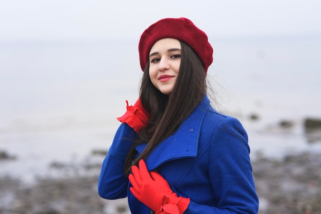 Portret pięknej dziewczyny rasy kaukaskiej, młoda atrakcyjna stylowa elegancka brunetka kobieta z długimi włosami w czerwonym berecie i rękawiczkach i niebieskim płaszczu, patrząc na kamery na zewnątrz, chodzenie na plaży, ocean.