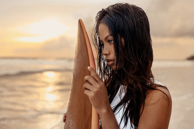 Portret pięknej dziewczyny rasy mieszanej surferów o mokrych włosach na plaży w profilu trzymającym białą deskę surfingową