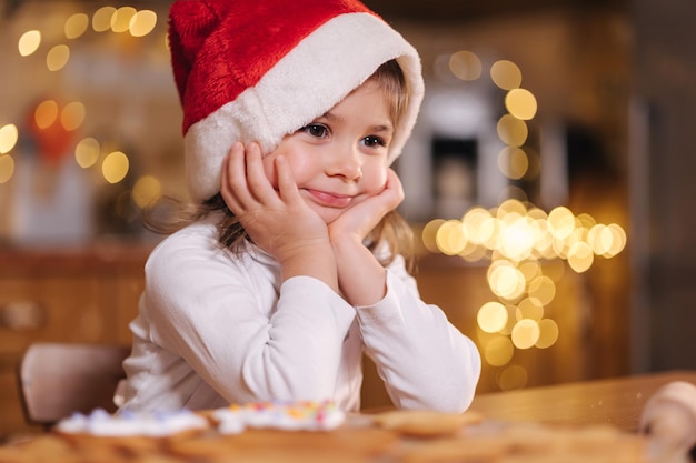 Portret pięknej dziewczyny roku drzewa w santa hat siedzi przy stole przed udekorowaną kuchnią