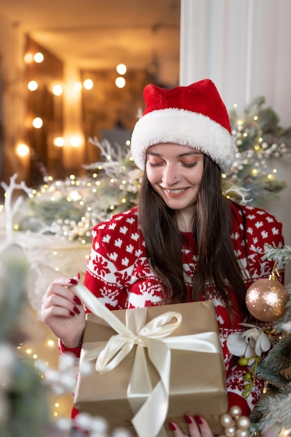 Portret pięknej dziewczyny w świątecznym swetrze i czerwonym kapeluszu Świętego Mikołaja stoi w pobliżu luksusowej choinki Uśmiecha się i trzyma prezent w dłoniach Obchody bożego narodzenia nowego roku