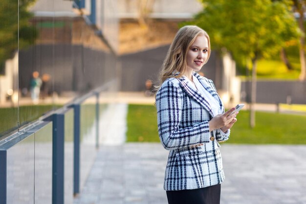 Portret pięknej eleganckiej bizneslady o blond włosach w kraciastej kurtce i spódnicy, trzymającej smartfon, wyglądającej pewnie i odnoszącej sukcesy, uśmiechającej się do kamery, stojącej w pobliżu centrum biznesowego na świeżym powietrzu