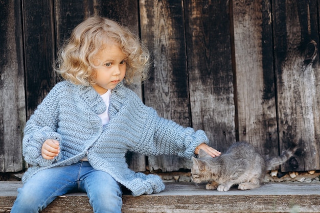 Zdjęcie portret pięknej i szczęśliwej kręconej dziewczyny w niebieskim swetrze z dzianiny bawi się z kotkiem w pobliżu drewnianej ściany